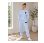 A woman modeling a light blue sweatshirt and jogger pants set with black heart details, standing in a bright room with a window and a potted plant.
