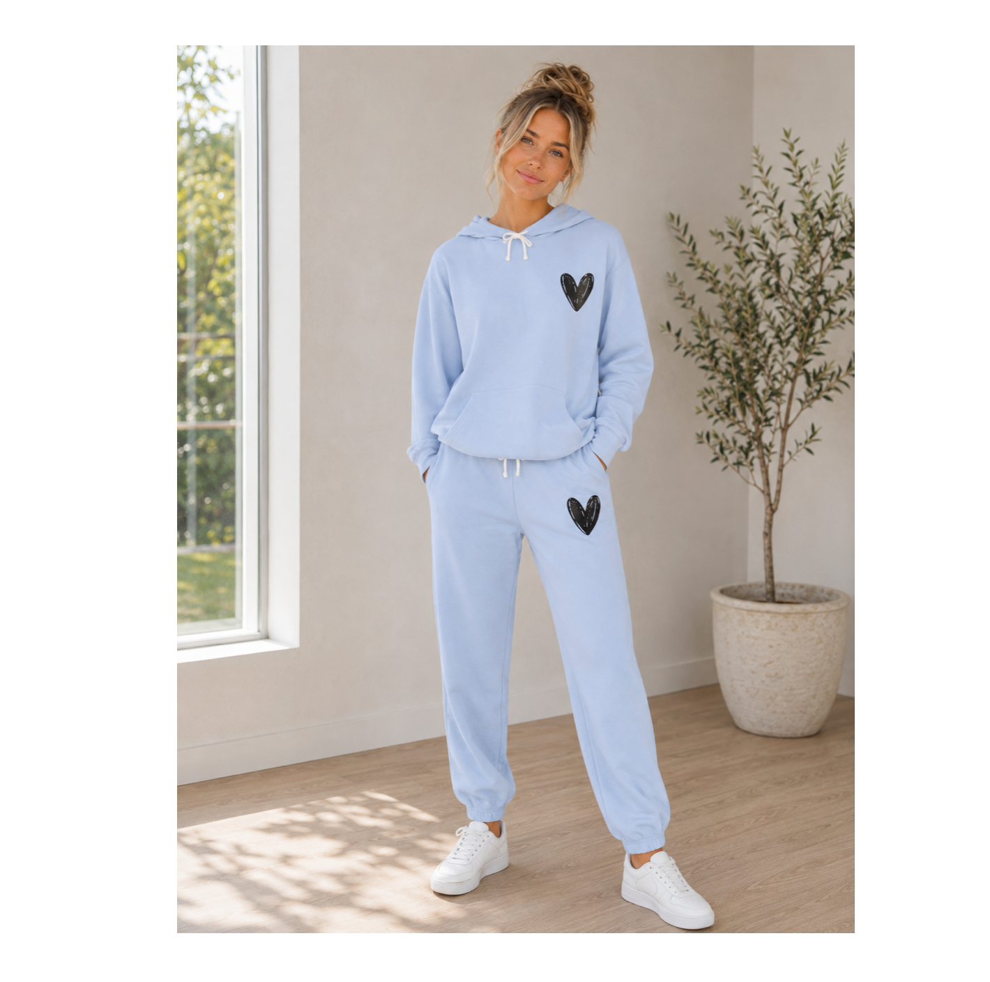 A woman modeling a light blue sweatshirt and jogger pants set with black heart details, standing in a bright room with a window and a potted plant.