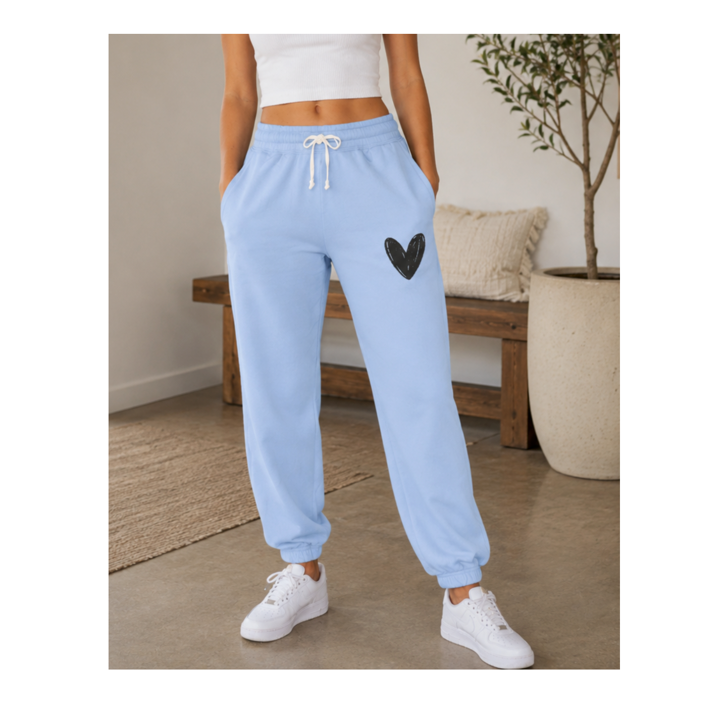 A person wearing light blue jogger sweatpants with a black heart patch on the thigh, paired with a white crop top and white sneakers, standing in a styled indoor room.