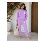 A woman modeling a lavender purple two-piece lounge set consisting of a pullover hoodie and matching jogger pants, both featuring a white heart graphic on the chest and thigh. She is holding a large woven straw tote bag and wearing white flip-flops, standing in front of a white wall with a wooden door and a potted plant.