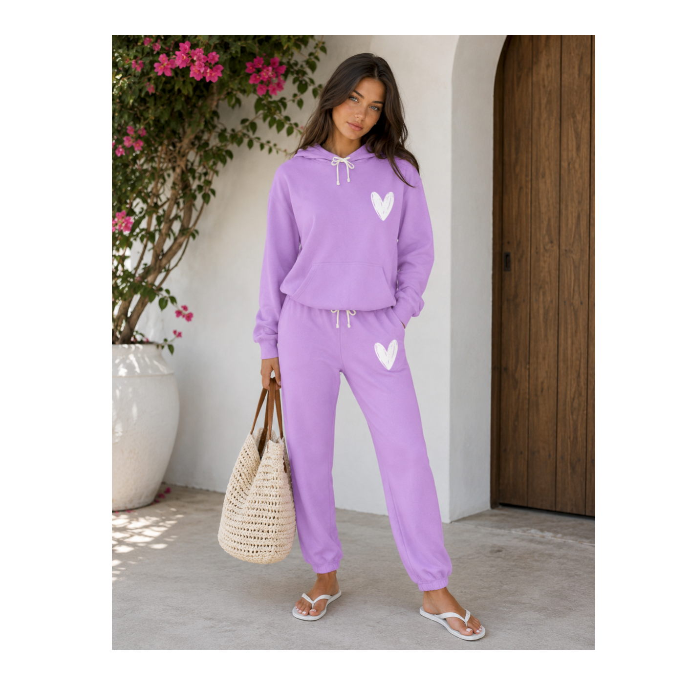 A woman modeling a lavender purple two-piece lounge set consisting of a pullover hoodie and matching jogger pants, both featuring a white heart graphic on the chest and thigh. She is holding a large woven straw tote bag and wearing white flip-flops, standing in front of a white wall with a wooden door and a potted plant.