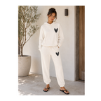A woman modeling a matching cream-colored lounge set consisting of a pullover hoodie and jogger pants, both featuring a black sequined heart on the chest and thigh. She is standing in a sunlit outdoor patio setting next to a potted olive tree.
