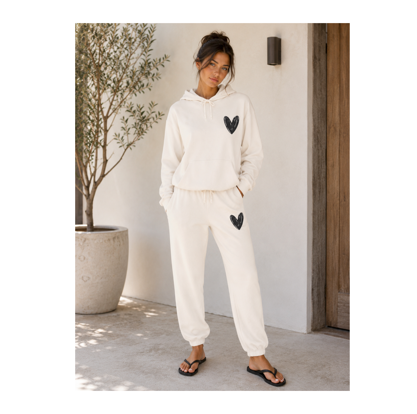 A woman modeling a matching cream-colored lounge set consisting of a pullover hoodie and jogger pants, both featuring a black sequined heart on the chest and thigh. She is standing in a sunlit outdoor patio setting next to a potted olive tree.