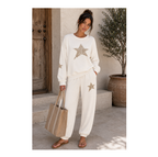 A woman stands outdoors wearing a matching cream lounge set with metallic gold star graphics on the sweatshirt and pants. She is holding a large beige tote bag and wearing brown sandals, set against a rustic background with a wooden door and a potted tree.
