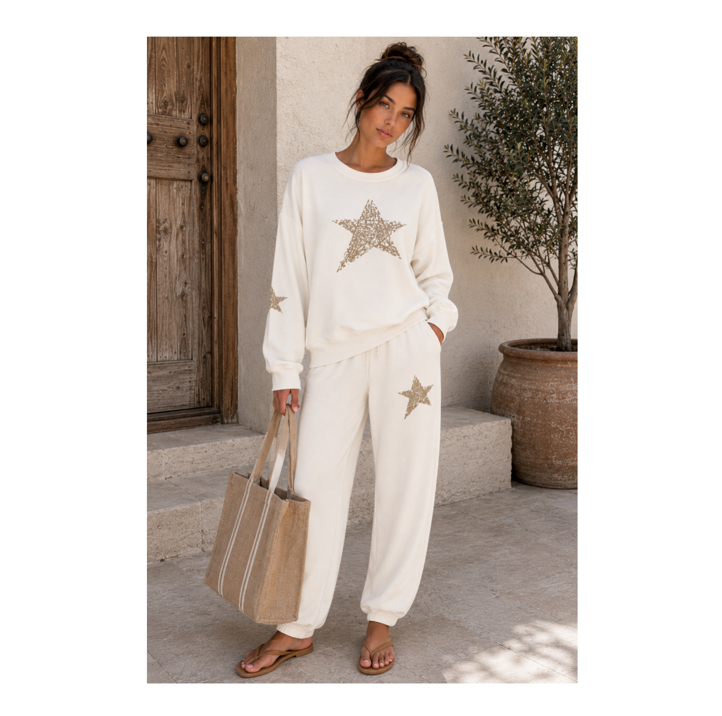 A woman stands outdoors wearing a matching cream lounge set with metallic gold star graphics on the sweatshirt and pants. She is holding a large beige tote bag and wearing brown sandals, set against a rustic background with a wooden door and a potted tree.