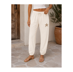 A person wearing cream-colored jogger sweatpants with a drawstring waist and a small star patch on the left thigh, standing on a stone patio next to a potted plant.
