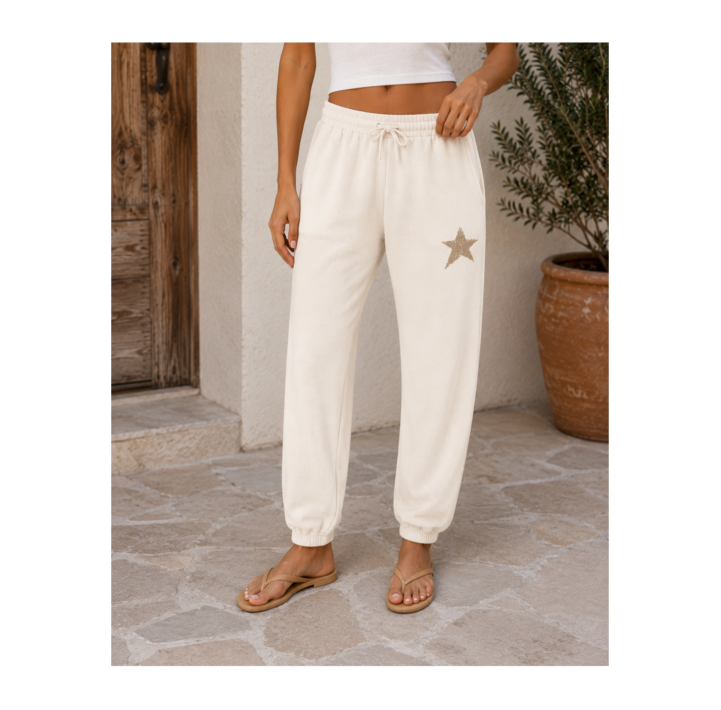 A person wearing cream-colored jogger sweatpants with a drawstring waist and a small star patch on the left thigh, standing on a stone patio next to a potted plant.