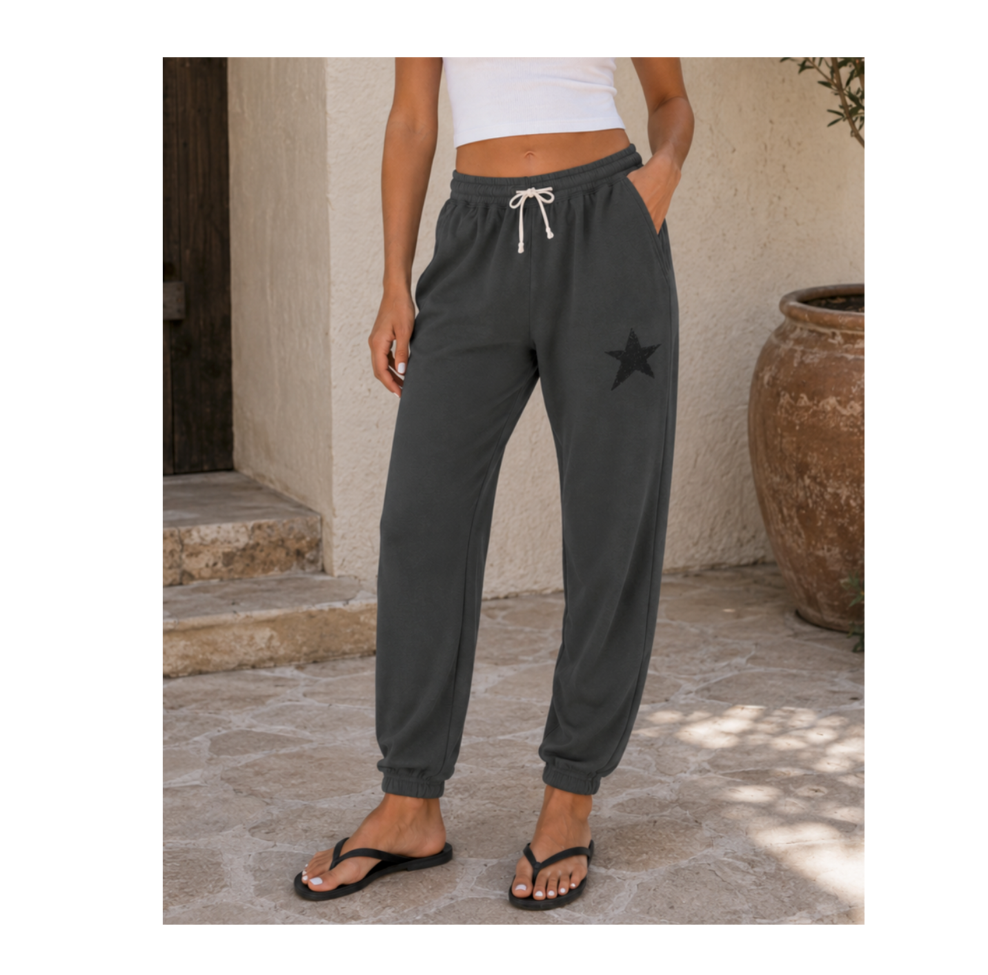 A person wearing dark charcoal gray jogger sweatpants with a black star graphic on the upper left thigh, white drawstrings, and elastic cuffs, standing outdoors on a stone patio.