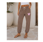 A person wearing brown jogger-style sweatpants with a white drawstring and a light pink heart graphic on the upper thigh, standing outdoors on a stone patio.