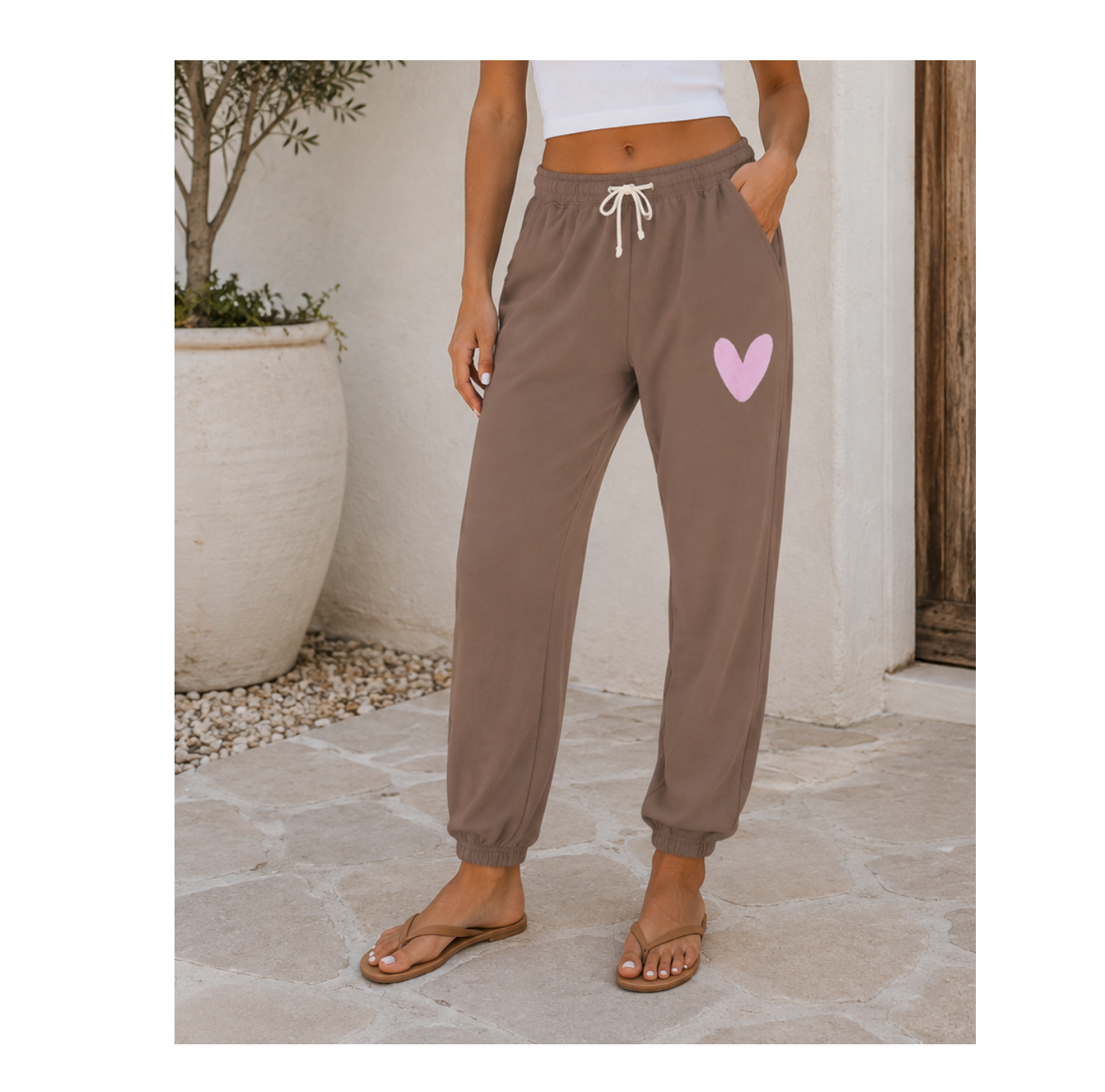 A person wearing brown jogger-style sweatpants with a white drawstring and a light pink heart graphic on the upper thigh, standing outdoors on a stone patio.