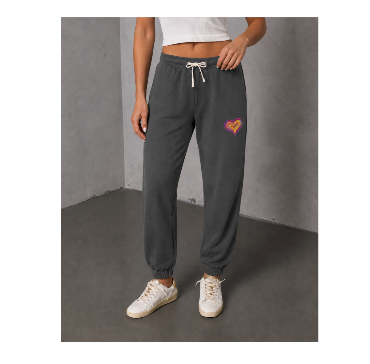 A person wearing dark gray jogger sweatpants with a small pink and orange heart graphic on the left thigh, paired with a white cropped tank top and white sneakers, standing against a gray concrete wall.