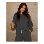 A woman modeling a dark gray sweatshirt and matching sweatpants set with red heart graphics, holding an iced coffee in front of a textured beige brick wall.