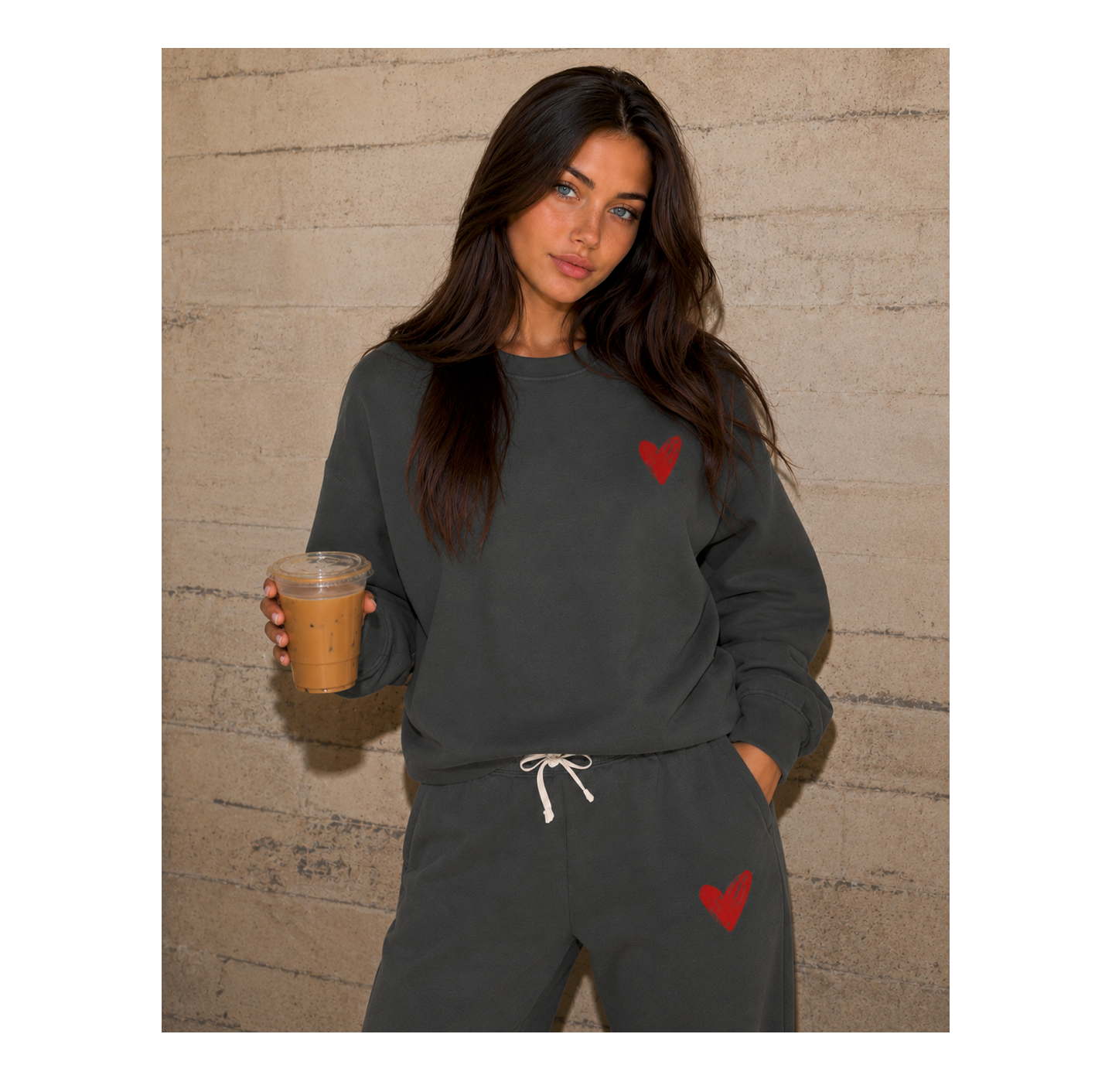 A woman modeling a dark gray sweatshirt and matching sweatpants set with red heart graphics, holding an iced coffee in front of a textured beige brick wall.